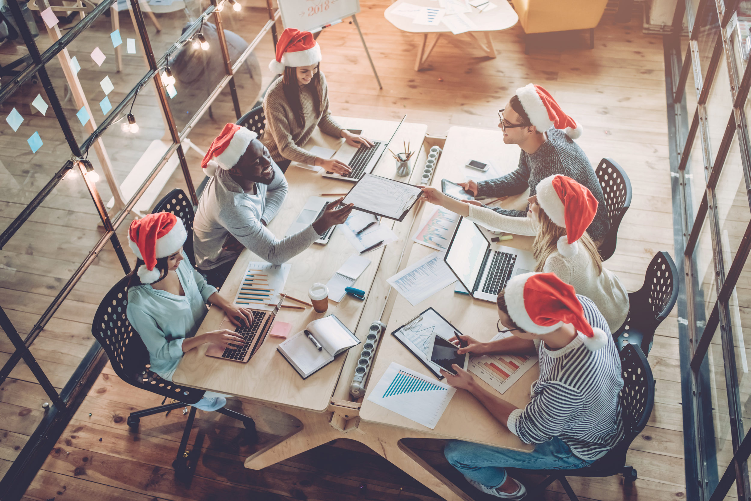 Merry Christmas and Happy New Year 2018! Multiracial young creative people are celebrating holiday in modern office. Group of young business people are sitting in Santa hats in last working day. Successful hipster team of freelancers.